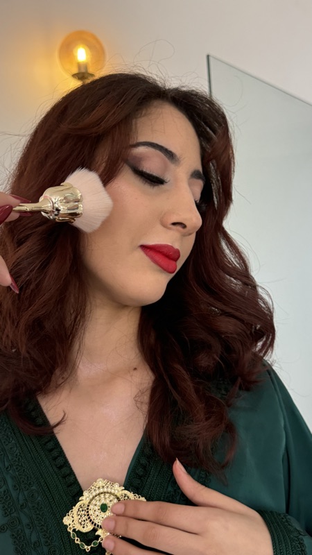 Close-up of matte red lipstick and warm sculpted cheek makeup on deeper olive skin, with a makeup brush in frame
